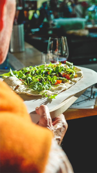 Veganer Flammkuchen mit Tomatensugo, Paprika, Oliven und Rucola, betr&auml;ufelt mit Oliven&ouml;l, serviert auf einem Holzbrett.