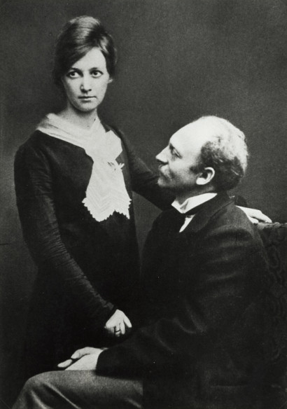 Ada Nolde st&aring;r i sort kjole med et hvidt t&oslash;rkl&aelig;de, Emil Nolde sidder p&aring; en stol i sort jakkes&aelig;t og ser p&aring; hende.