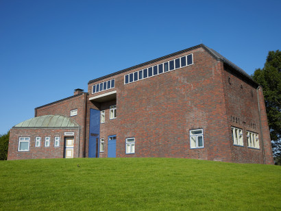 Exterior view of the Nolde House with a blue entrance door, red brickwork, and the distinctive window front of the picture gallery; the building is reminiscent of the Bauhaus style of the 1920s.